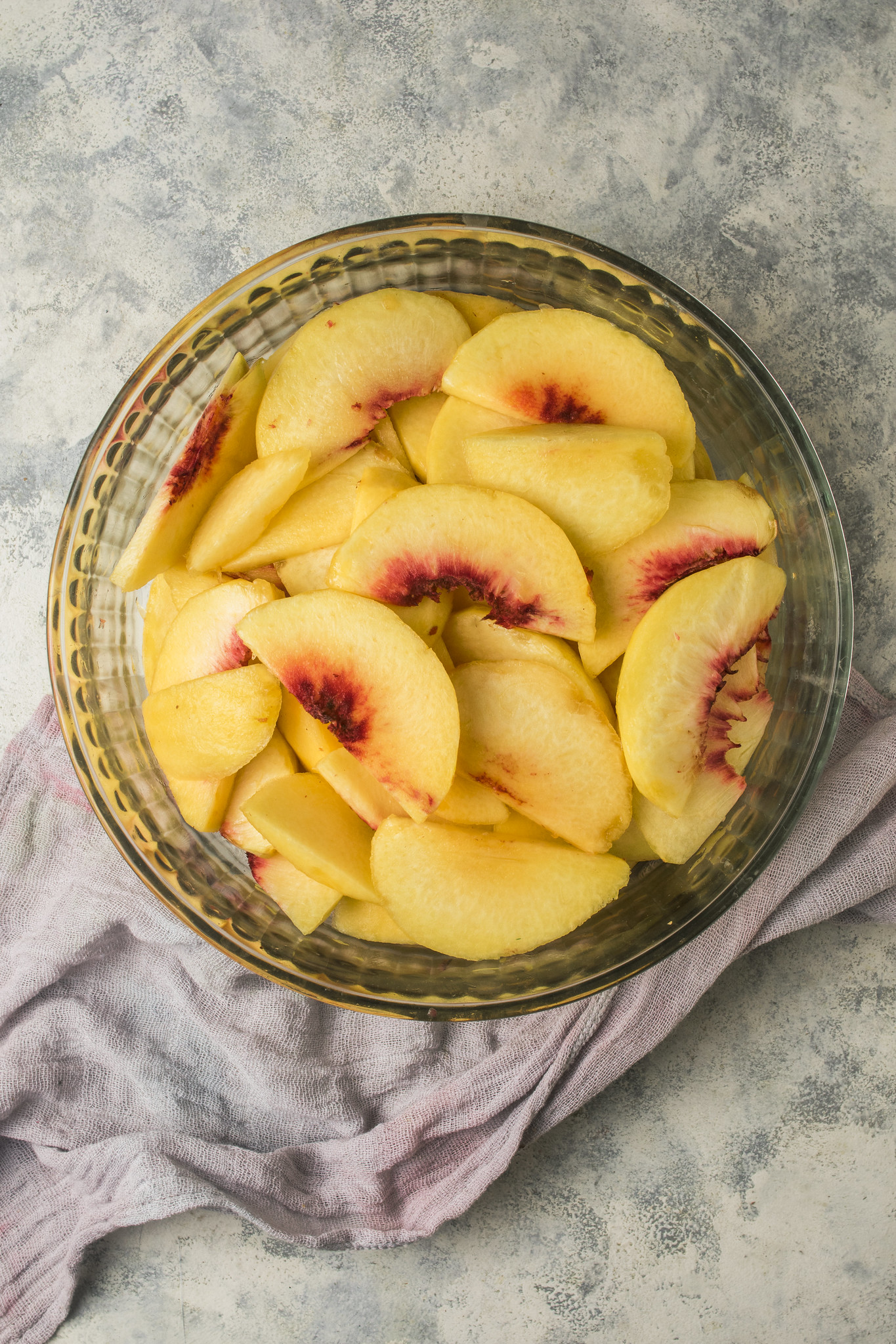 Overhead view of sliced fresh peaches in a glass bowl, prepared for sbriciolata di pesche, on a softly textured surface with a pale cloth.